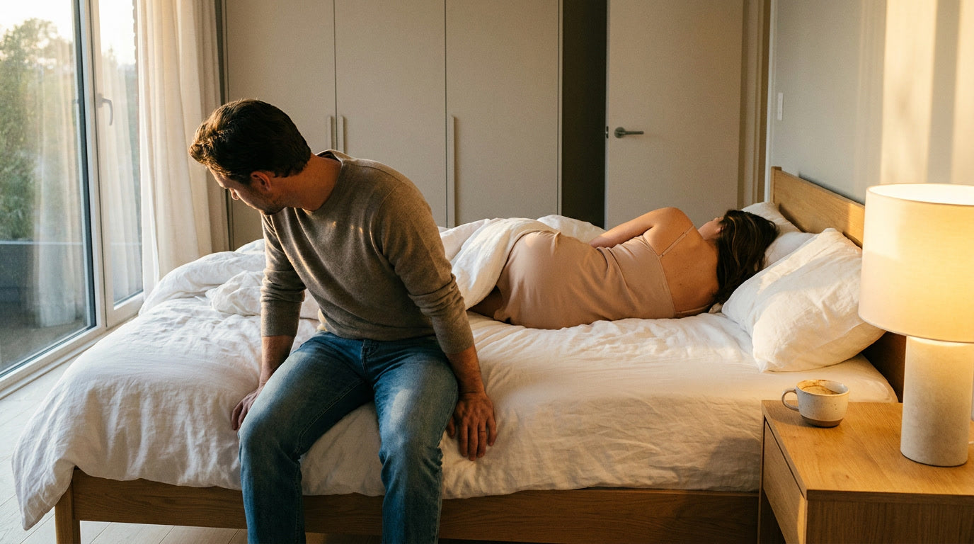 Homme assis au bord du lit regardant par la fenêtre, femme tournée de dos dans les draps froissés, distance silencieuse entre
