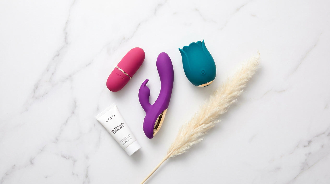 Trois vibromasseurs clitoridiens colorés rose violet teal posés sur marbre blanc avec lubrifiant et fleur séchée
