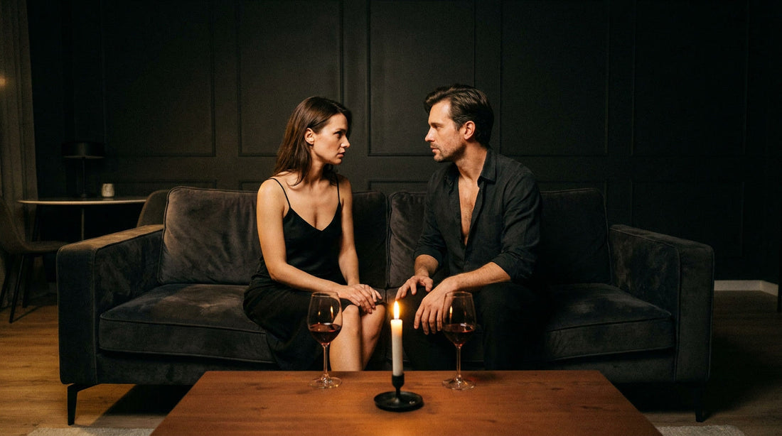 Couple assis face à face sur un canapé sombre, regards intenses, deux verres de vin sur la table, conversation intime sur le 