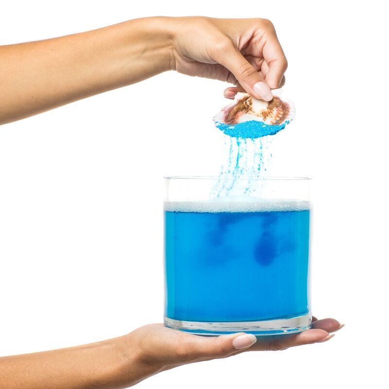Main versant les sels de bain Kamasutra Treasures of the Sea depuis un coquillage dans un verre d'eau qui se colore en bleu, demonstration de l'effet apaisant ocean.