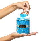 Mains tenant le pot Kamasutra Treasures of the Sea, sels de bain de luxe aux mineraux marins, demonstration du contenu bleu apaisant et adoucissant pour la peau.