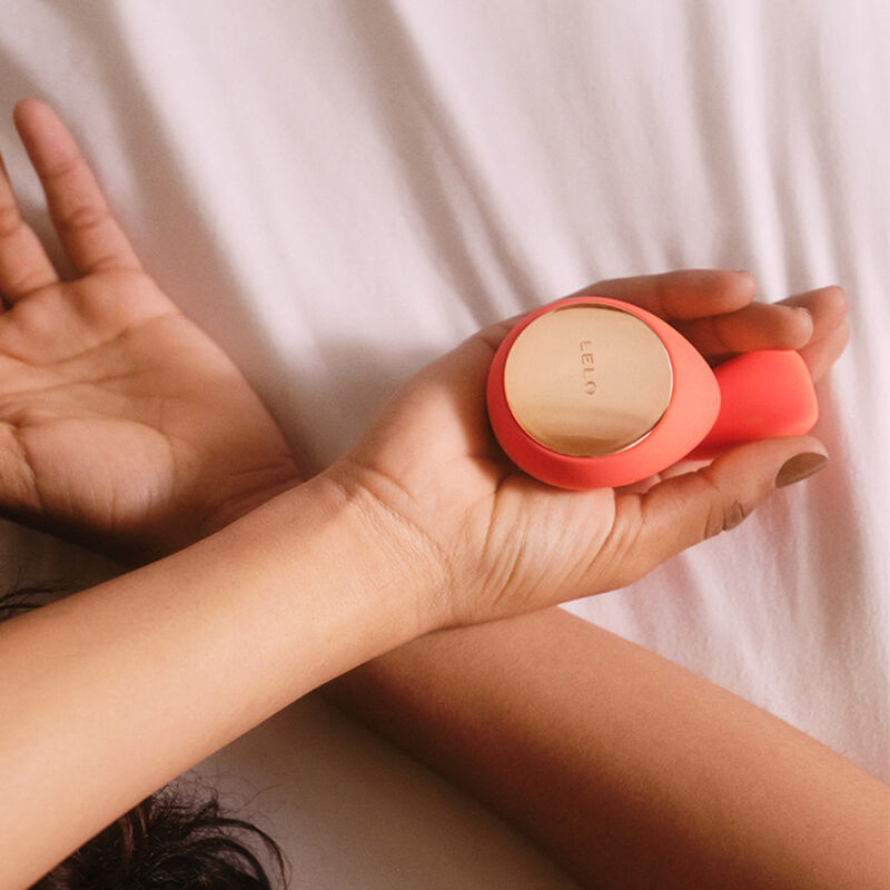 Main tenant le stimulateur de couple IDA Wave de Lelo en silicone corail, gros plan sur sa surface lisse et sa finition dorée posée sur un drap clair.