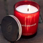 Gros plan sur la bougie de massage rouge Matchmaker allumee, couvercle ouvert posant a cote, cire fondue du soin aux pheromones 150 ml Eye Of Love.
