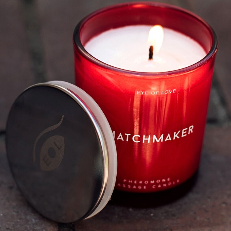 Gros plan sur la bougie de massage rouge Matchmaker allumee, couvercle ouvert posant a cote, cire fondue du soin aux pheromones 150 ml Eye Of Love.