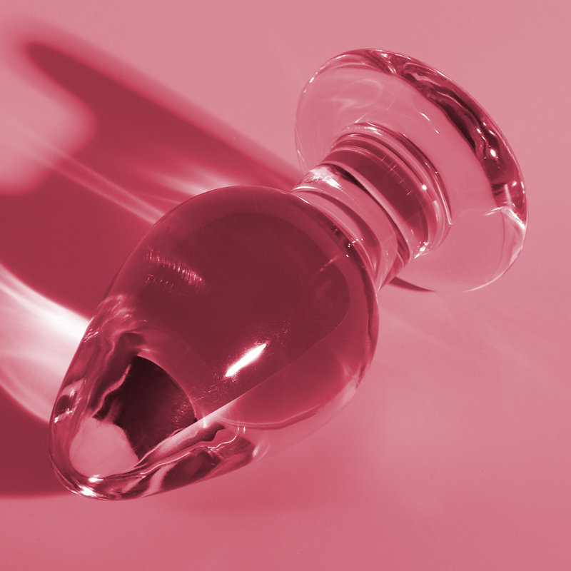 Gros plan en biais du plug anal conique en verre borosilicate sur fond rose, base annelee en avant-plan et corps bombe lisse, surface non poreuse hygienique.