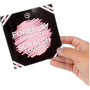 Jeu de grattage Foreplay Fanatics Scratch Lovers tenu en main, carte noire a cases roses. Gratte une option par categorie pour creer une combinaison de preliminaires coquins.