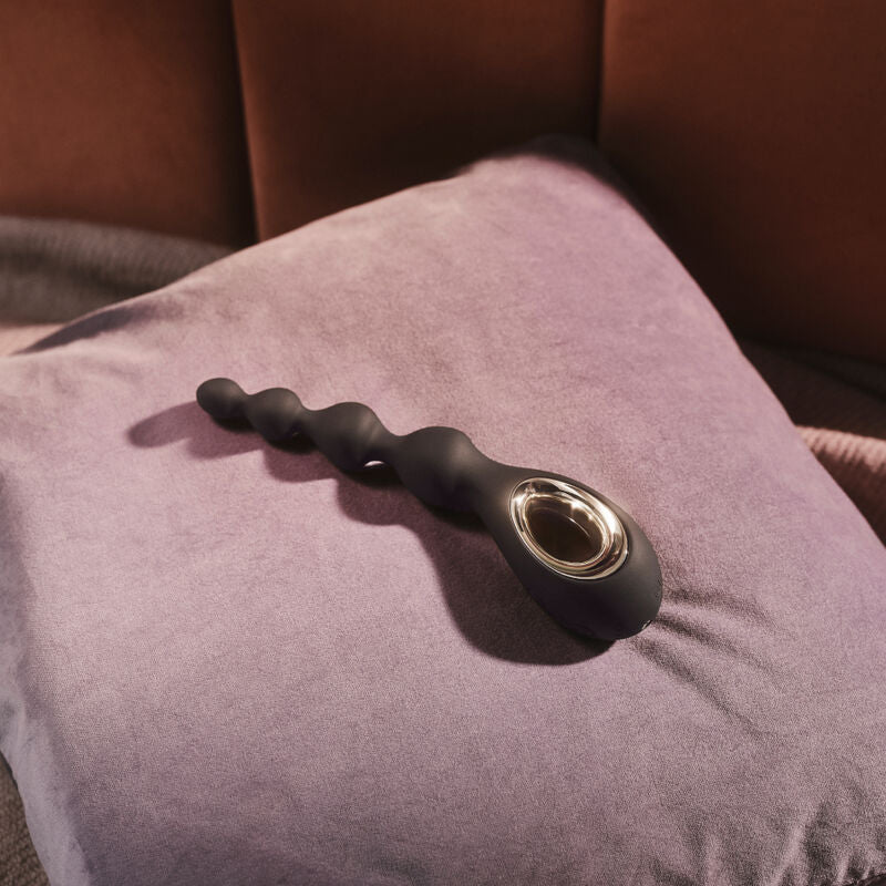 Chapelet anal Soraya Beads noir de Lelo pose sur un coussin rose, mise en scene lifestyle d'un sextoy a perles en silicone doux a poignee doree vibrant a pulsation