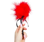 Plumeau tickler Secret Play en plumes rouges tenu en main par le manche noir a ruban, accessoire de caresse sensorielle pour eveiller la peau et pimenter les preliminaires.