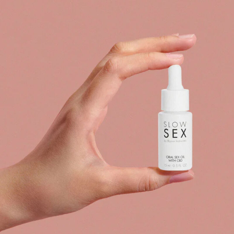 Main tenant le flacon compte-gouttes Oral Sex Oil with CBD Slow Sex Bijoux Indiscrets entre les doigts sur fond rose, format compact de l'huile gourmande 15 ml