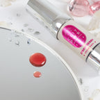 Flacon argente du baume Clit Pwr fraise pose pres d'une goutte rose sur un miroir, baume stimulant clitoridien chauffant et picotant Secret Play, texture fluide.