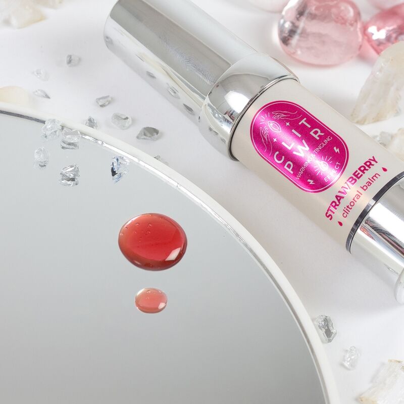 Flacon argente du baume Clit Pwr fraise pose pres d'une goutte rose sur un miroir, baume stimulant clitoridien chauffant et picotant Secret Play, texture fluide.