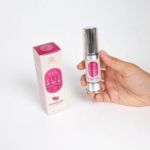 Flacon airless de baume Clit Pwr fraise tenu en main devant sa boite rose Secret Play, baume clitoridien stimulant effet chaud et picotant, absorption rapide externe.