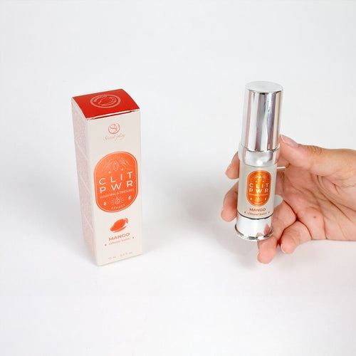 Flacon airless du baume clitoris Clit Pwr mangue tenu en main pres de sa boite orange. Soin stimulant externe a sensation de chaleur et picotement, saveur mangue gourmande.