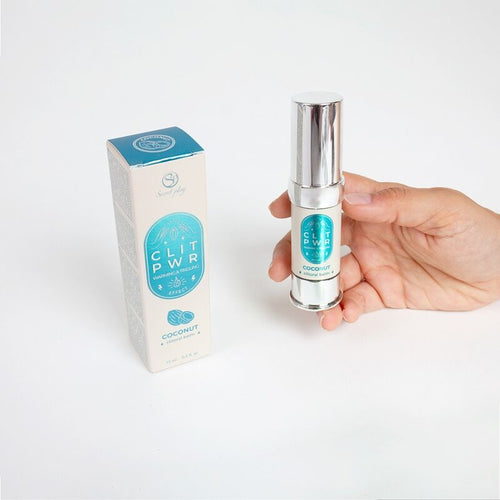 Flacon airless du baume clitoris Clit Pwr noix de coco tenu en main pres de sa boite bleue. Soin stimulant externe a sensation de chaleur et picotement, texture non collante.