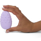 Masseur Mimi Deux lilas tenu du bout des doigts d'une main, galet texturé de picots vu de profil, plan présentant le format de poche et la prise du produit sur fond blanc épuré.