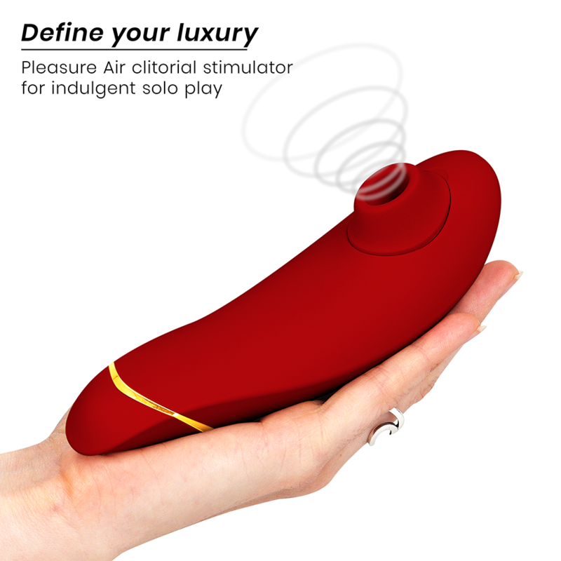 Main ouverte présentant le Womanizer Premium 2 rouge au liseré doré posé à plat, embout d'air pulsé vers la gauche, schéma d'ondes d'air clitoridien. Format ergonomique compact.