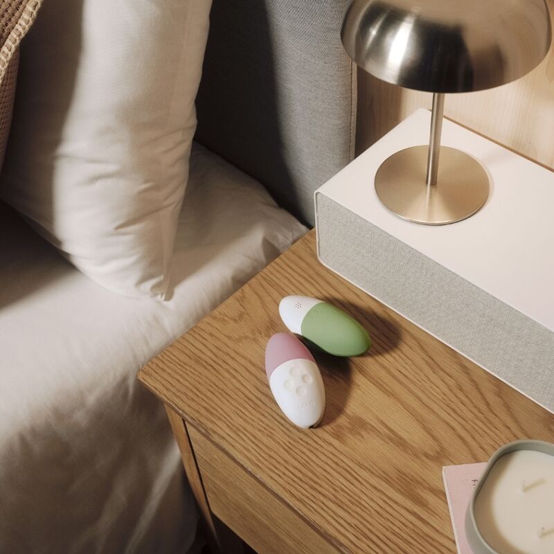 Gros plan de deux stimulateurs Lelo Siri 3 vert et rose sur table de chevet en bois pres d'une lampe doree, galets vibrants en silicone actives par le son, decor cosy.