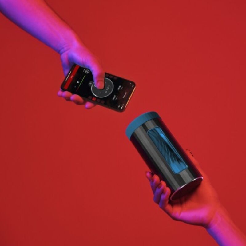 Masturbateur Lelo F1S V3 tenu en main face a un smartphone sur fond rouge, console de plaisir connectee pilotee par appli sans anatomie visible.