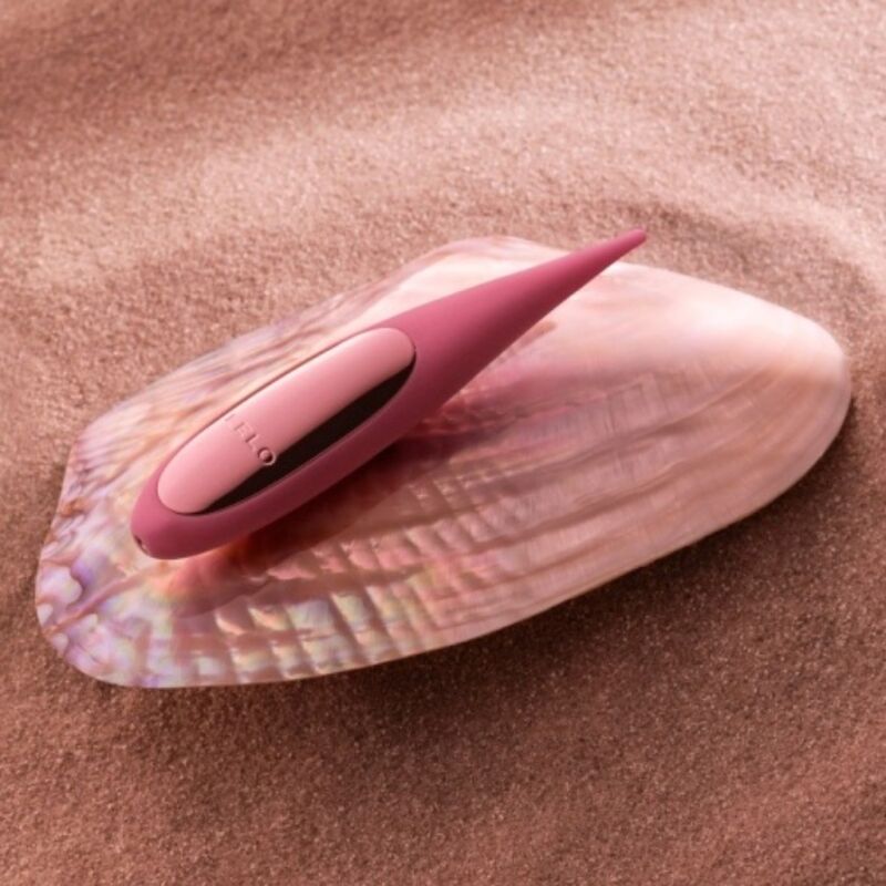 Vibromasseur de précision Dot rose posé sur un coquillage nacré dans le sable, mise en scène esthétique de l'embout pointu souple en silicone, ambiance balnéaire.