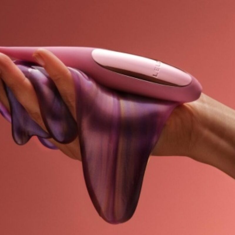 Vibromasseur de précision Dot rose tenu contre une étoffe violette drapée sur une main, mise en scène esthétique de l'embout pointu souple, fond corail.