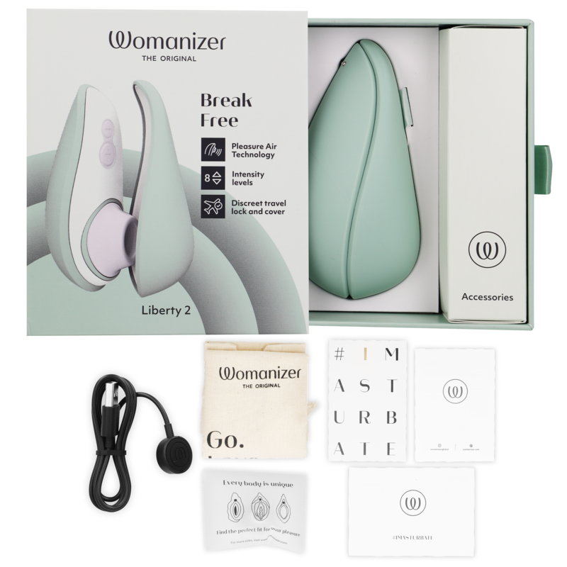 Coffret du Womanizer Liberty 2 vert sauge ouvert sur fond beige avec stimulateur, câble USB, pochette et cartes d'accessoires, packaging Break Free disposé autour.