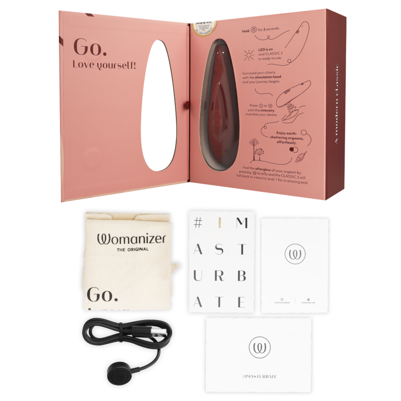 Coffret rose ouvert du Womanizer Classic 2, câble de charge USB, sachet de rangement et cartes de notice présentés à plat, stimulateur clitoridien rechargeable.