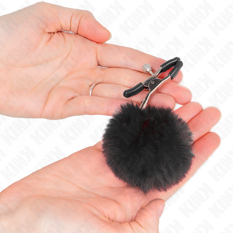Pince a tetons KINK a pompon en fausse fourrure noire posee dans la paume, gros plan sur le contraste entre la machoire metal et la touffe moelleuse.