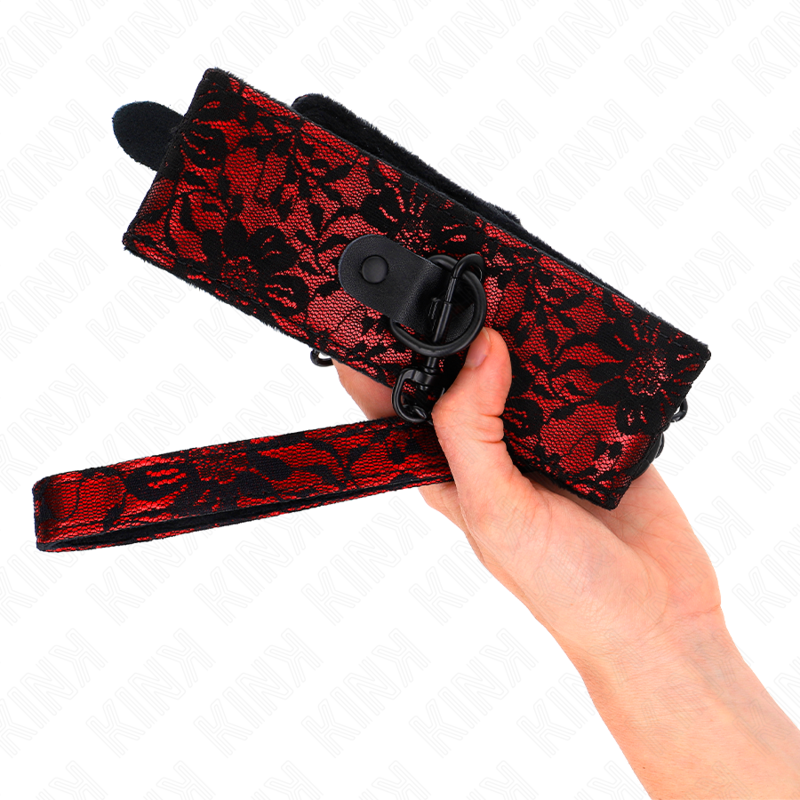 Collier de soumission a motif dentelle rouge et noir tenu en main, sangle a anneaux et boutons-pression, accessoire fetiche chic du jeu sub a deux.
