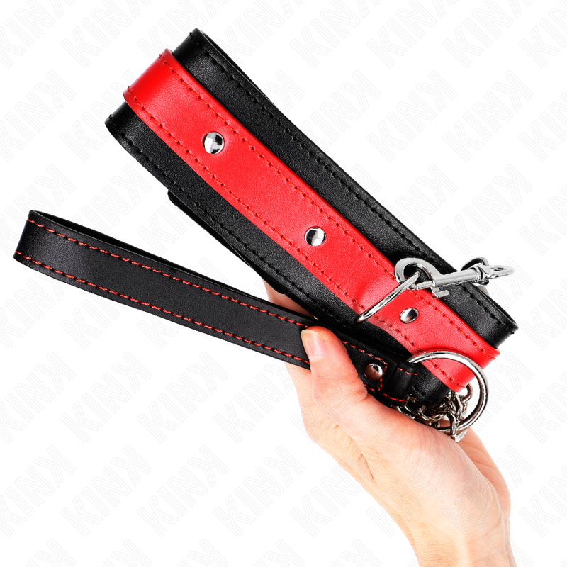 Collier de soumission rouge et noir tenu en main, anneaux argentés et coutures rouges visibles. Détail du collier-laisse BDSM bicolore.