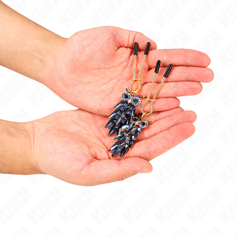 Pinces à tétons à breloques hibou bleu tenues au creux des mains, gros plan sur les pendentifs émaillés, accessoire de stimulation sensorielle élégant.