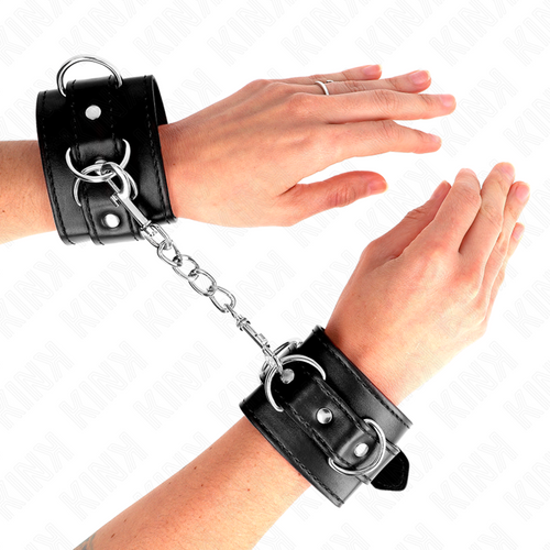 Manchettes de poignets en cuir noir portees aux poignets croises, mains ouvertes, chaine de liaison entre les deux, contention BDSM en cuir veritable.