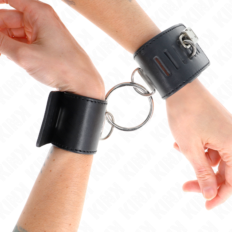 Larges menottes de poignets en cuir noir reliees par un grand anneau metallique central, aux poignets : contention rapprochee pour les jeux de soumission BDSM.
