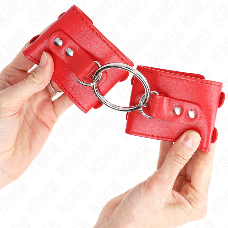 Manchettes de poignets rouges à anneau d'attache présentées à deux mains, détail des rivets et de l'anneau central de l'accessoire Kink.