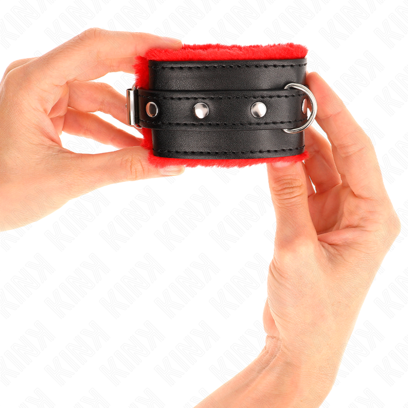 Manchette de poignet en cuir noir bordee de fourrure rouge, tenue entre deux mains pour montrer la sangle a boutons-pression et l'interieur moelleux de cette entrave douce.
