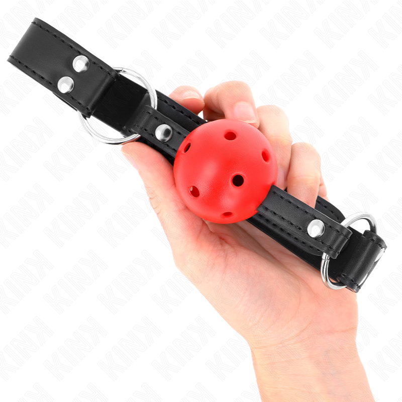 Bâillon boule rouge perforée tenu en main, sangle noire et anneaux métalliques visibles de chaque côté de la boule.