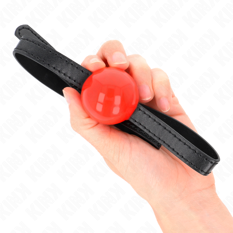 Bâillon boule rouge sur lanière tenu en main, échelle visible de la boule brillante et de la sangle noire, accessoire de soumission BDSM compact et coloré.