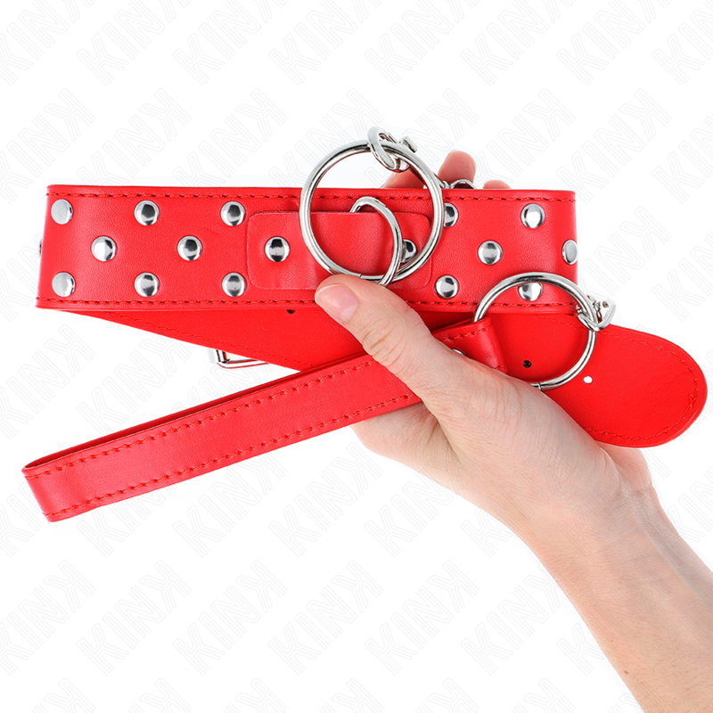 Collier de soumission rouge clouté tenu en main, gros plan sur l'anneau central et les rivets, accessoire BDSM dominant-soumis coloré et solide.