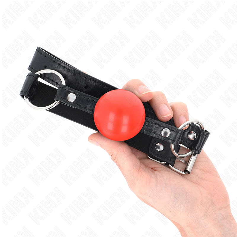 Bâillon boule rouge en caoutchouc tenu en main avec sa boucle métallique, échelle visible de cet accessoire de soumission BDSM de 4 cm sur sangle à anneaux.