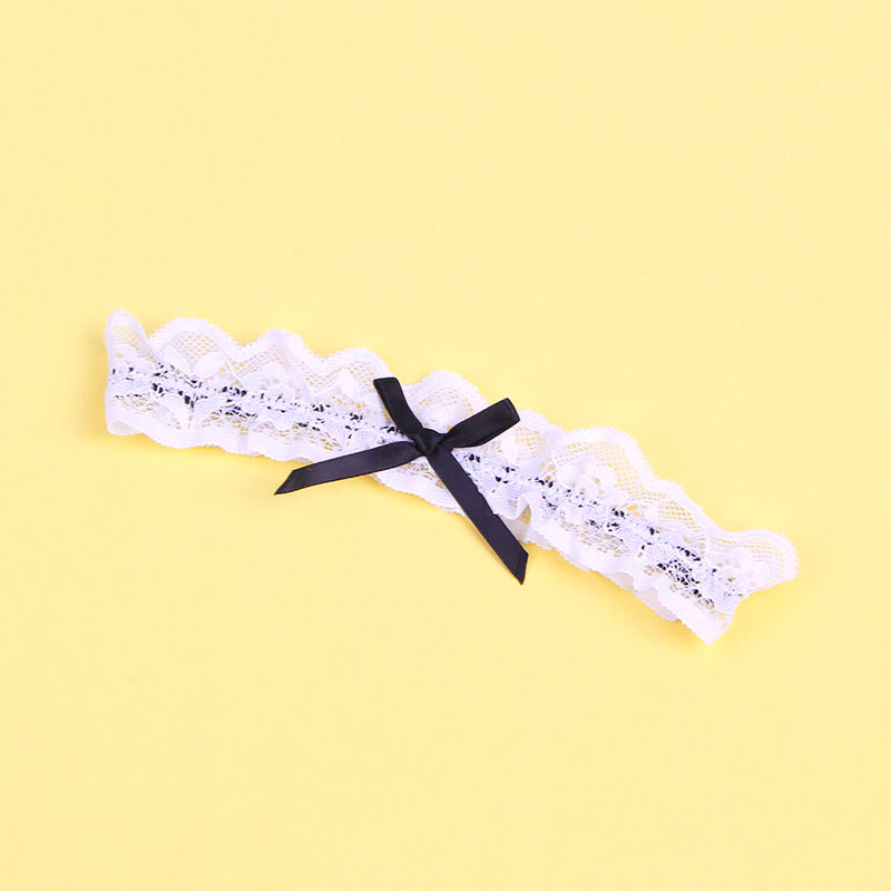 Gros plan a plat de la jarretiere en dentelle blanche du costume soubrette Subblime ornee d'un noeud noir central, presentee seule etalee sur un fond jaune uni lumineux.