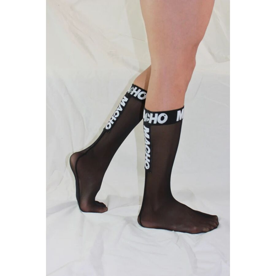 Chaussette haute homme noire semi-transparente portee de profil, bord logo Macho blanc, maille fine fetichiste soulignant le galbe du mollet.