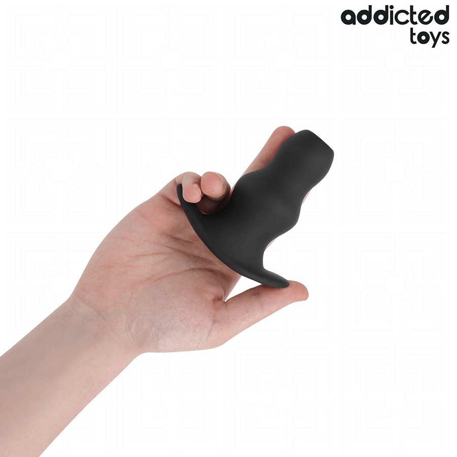 Plug anal creux Addicted en silicone noir tenu en main, tunnel central visible et base large profilee, format pratique pour decouvrir la stimulation par ouverture anale.