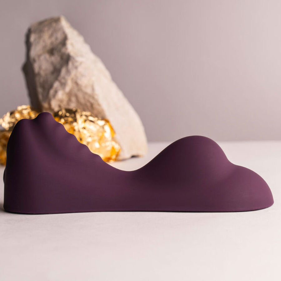 Vibromasseur à chevaucher violet en forme de selle posé devant une pierre grise et un éclat doré, double bosse ondulée lisse présentée sur fond clair neutre et épuré.