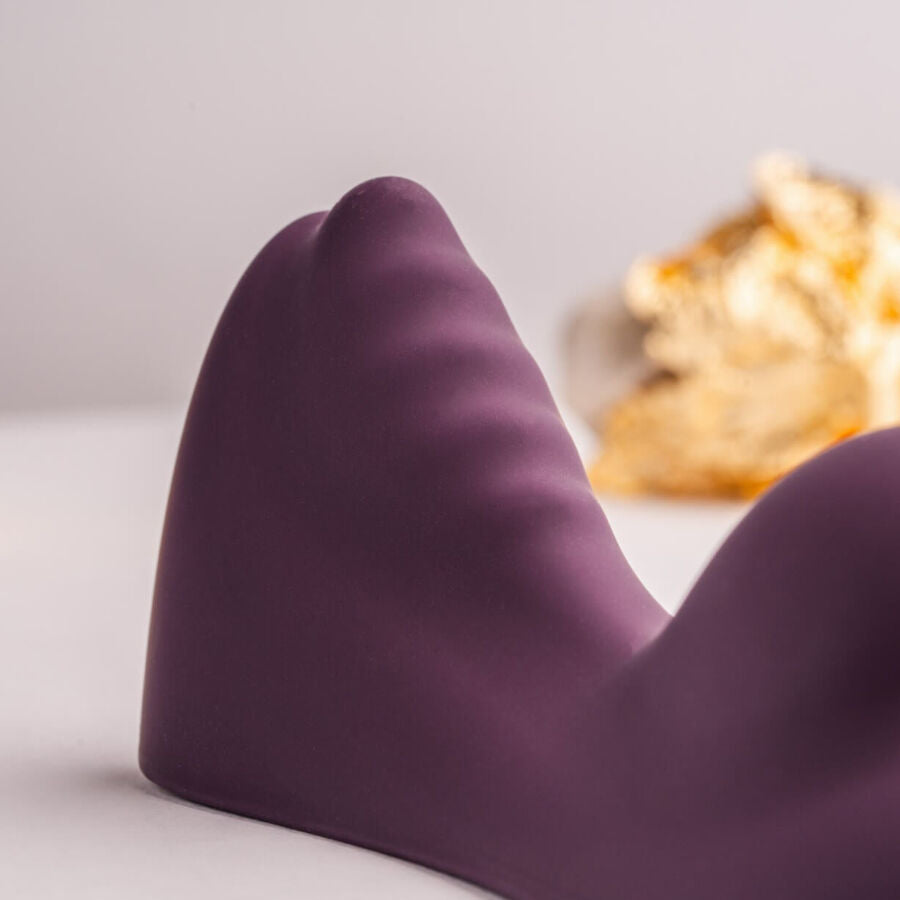 Gros plan du vibromasseur à chevaucher violet montrant ses deux bosses et les nervures de surface, éclat doré flouté en fond, silicone mat lisse sur fond clair neutre.