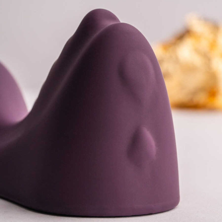 Macro du relief central du vibro à chevaucher violet aux petits picots, courbes douces en silicone mat, arrière-plan doré flouté sur fond clair neutre et sobre.