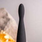 Plug anal Teazer noir effilé dressé devant une pierre, tige souple en silicone et base ergonomique, stimulateur point P fin 7 niveaux étanche signé Rocks-Off.