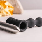 Composants du plug perlé posés ensemble : manchon silicone noir et bullet argentée gravée du logo, surfaces lisses sur fond clair sans contexte corporel.