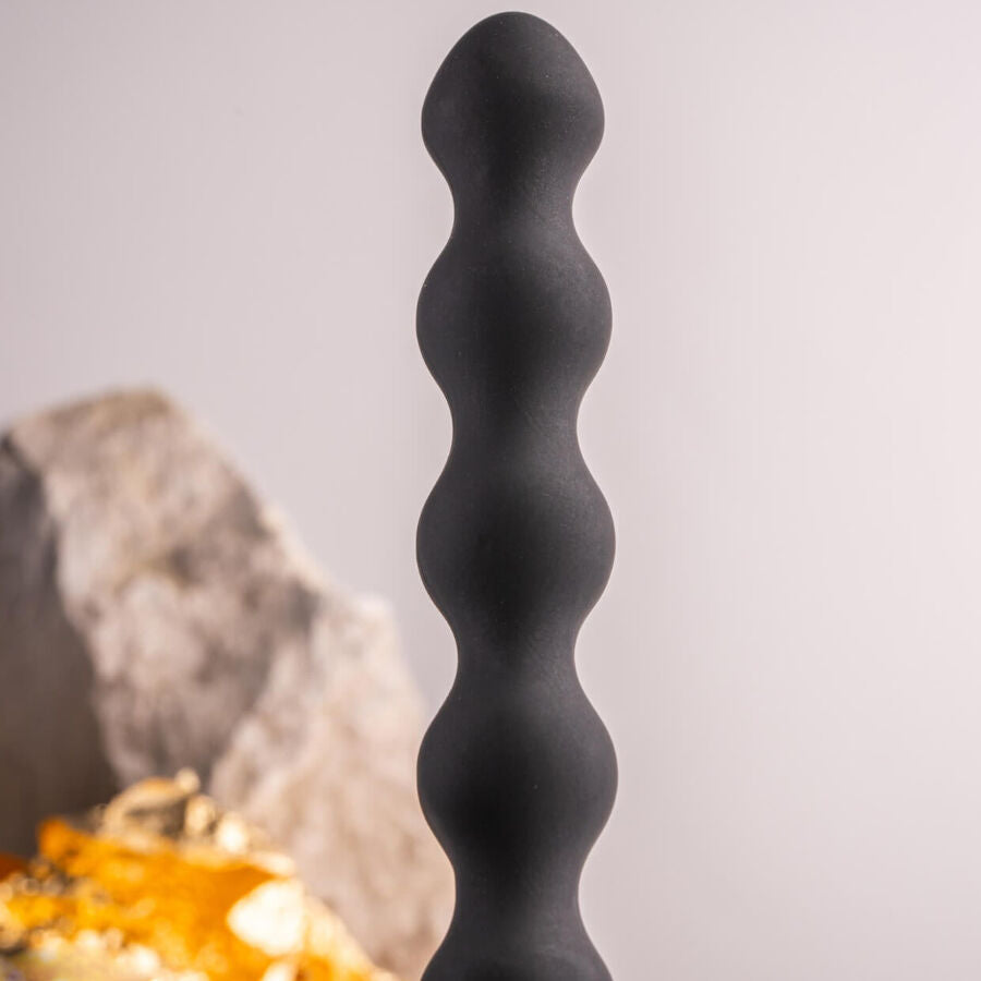 Plug anal perlé noir dressé près de pierres claires et d'un éclat doré, billes étagées et col fin lisses, silicone mat présenté sur fond neutre épuré et sobre.
