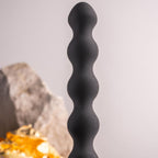 Plug anal perlé noir dressé près de pierres claires et d'un éclat doré, billes étagées et col fin lisses, silicone mat présenté sur fond neutre épuré et sobre.