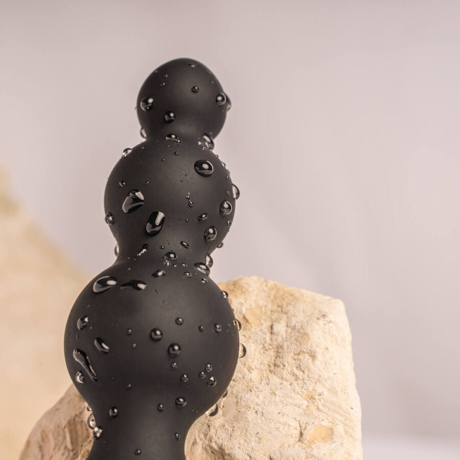Macro du plug à billes noires couvert de gouttes d'eau appuyé sur une pierre claire, renflements étagés lisses illustrant l'étanchéité de l'accessoire.