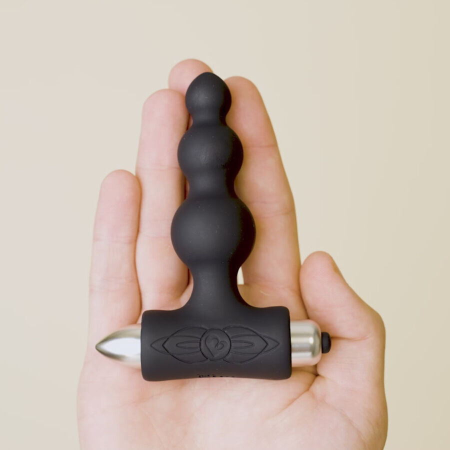Plug anal à billes noir tenu au bout des doigts illustrant son petit format, base vibrante en T à embout métallisé visible, silicone lisse sur fond neutre.